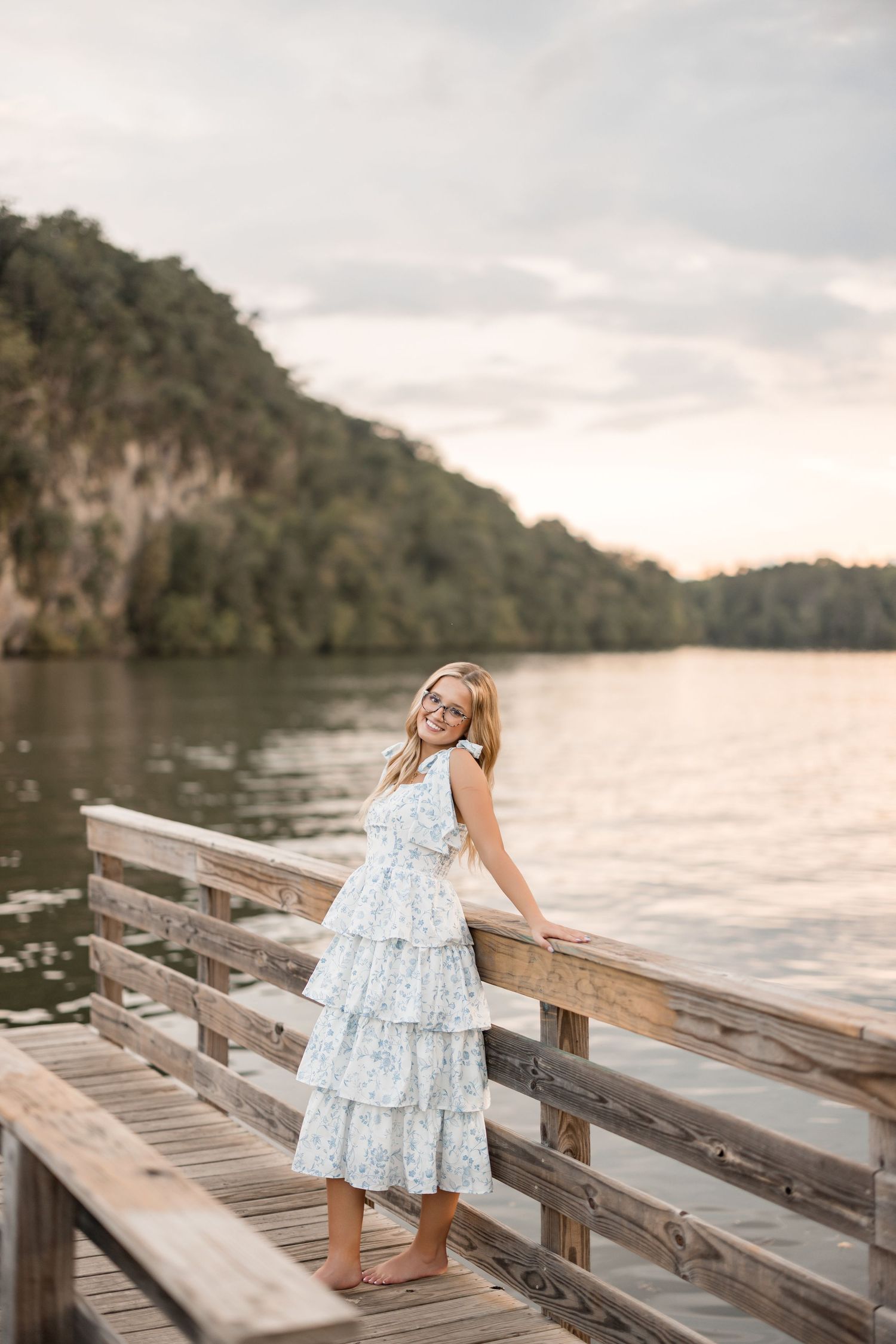 A small child in a blue and white dress walks along a wooden dock by a peaceful lake at sunset.