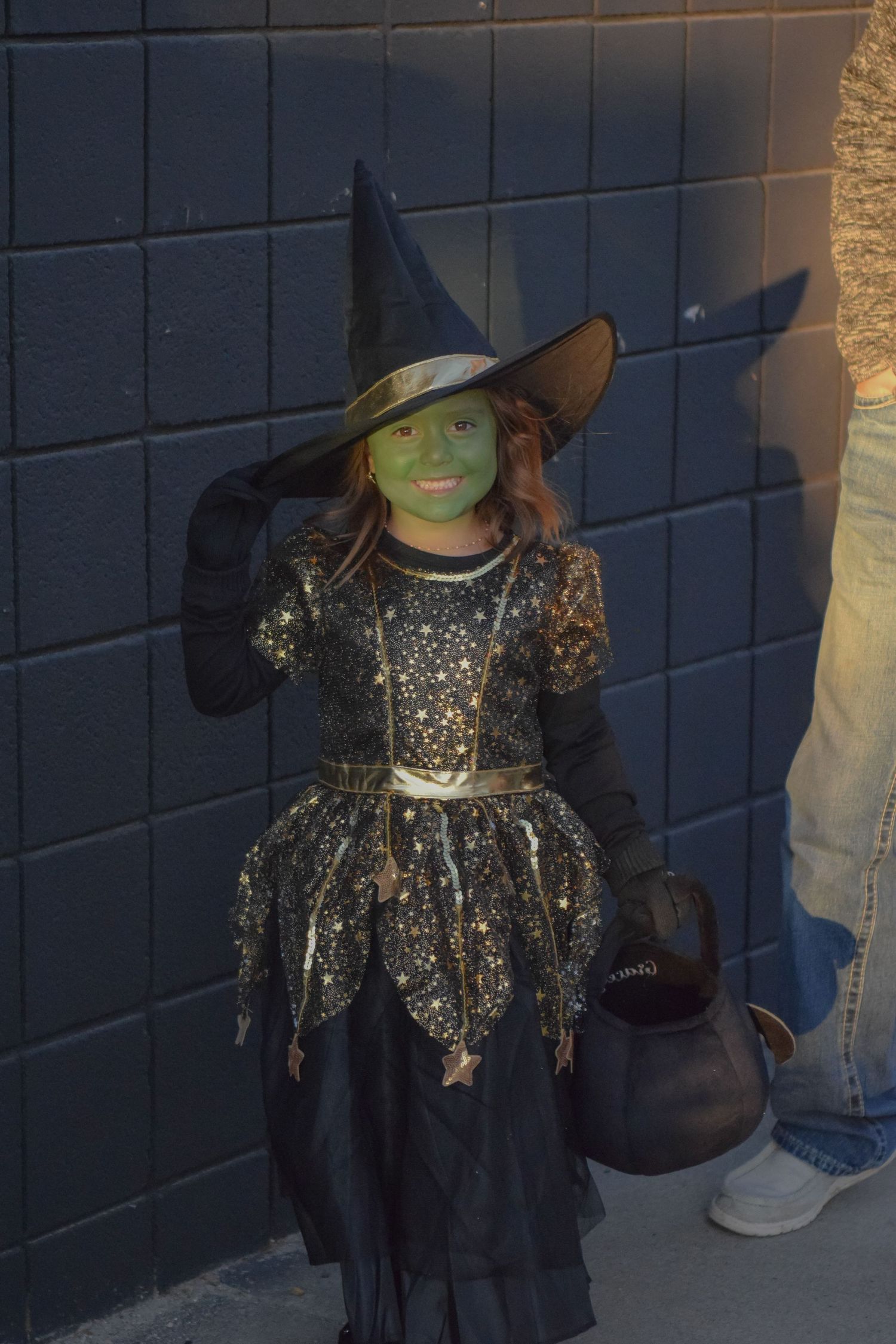 A figure in a sparkly witch costume with green face paint and black hat poses against a dark wall.