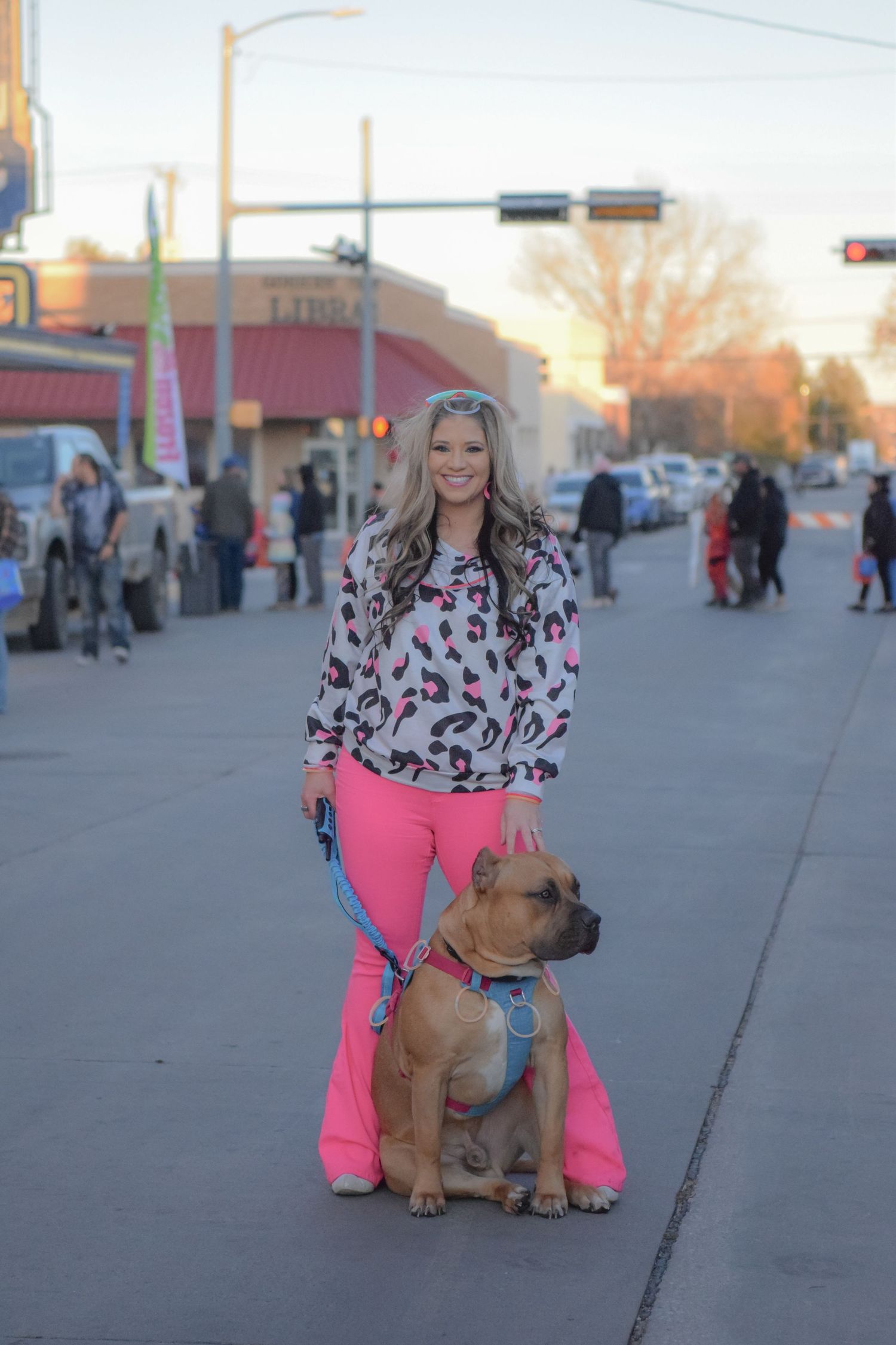 A person in pink pants poses with a brown pit bull dog on a sidewalk.
