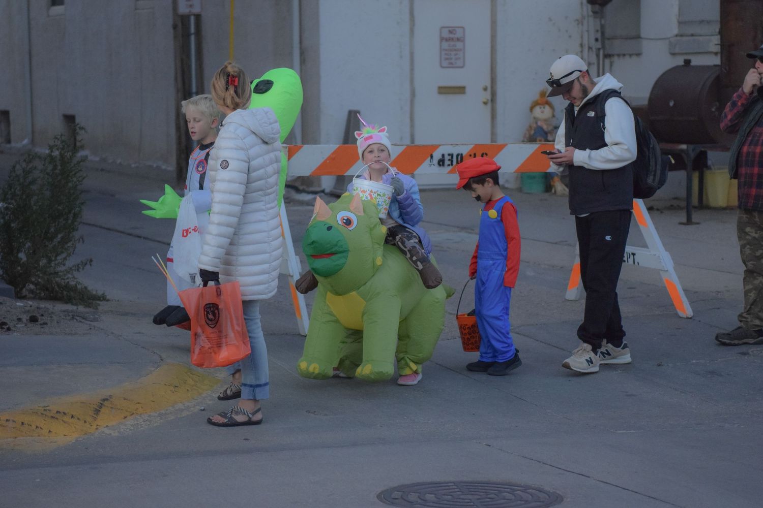 Children in colorful Halloween costumes including dinosaur and superhero outfits walk down a sidewalk trick-or-treating.