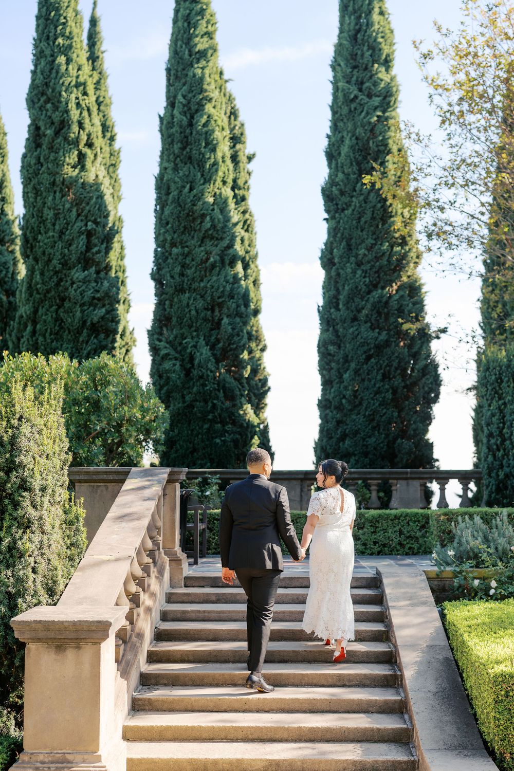 A couple ascends elegant stone steps flanked by tall cypress trees in a formal garden setting.