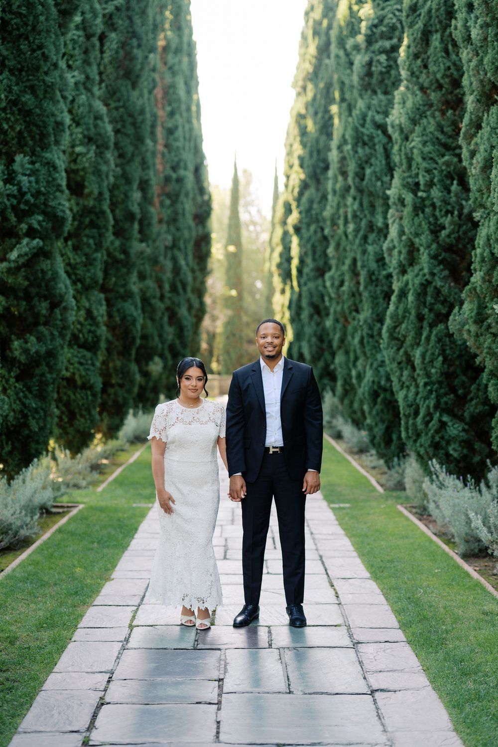 A couple poses playfully on a stone path lined with tall cypress trees in a romantic garden setting.