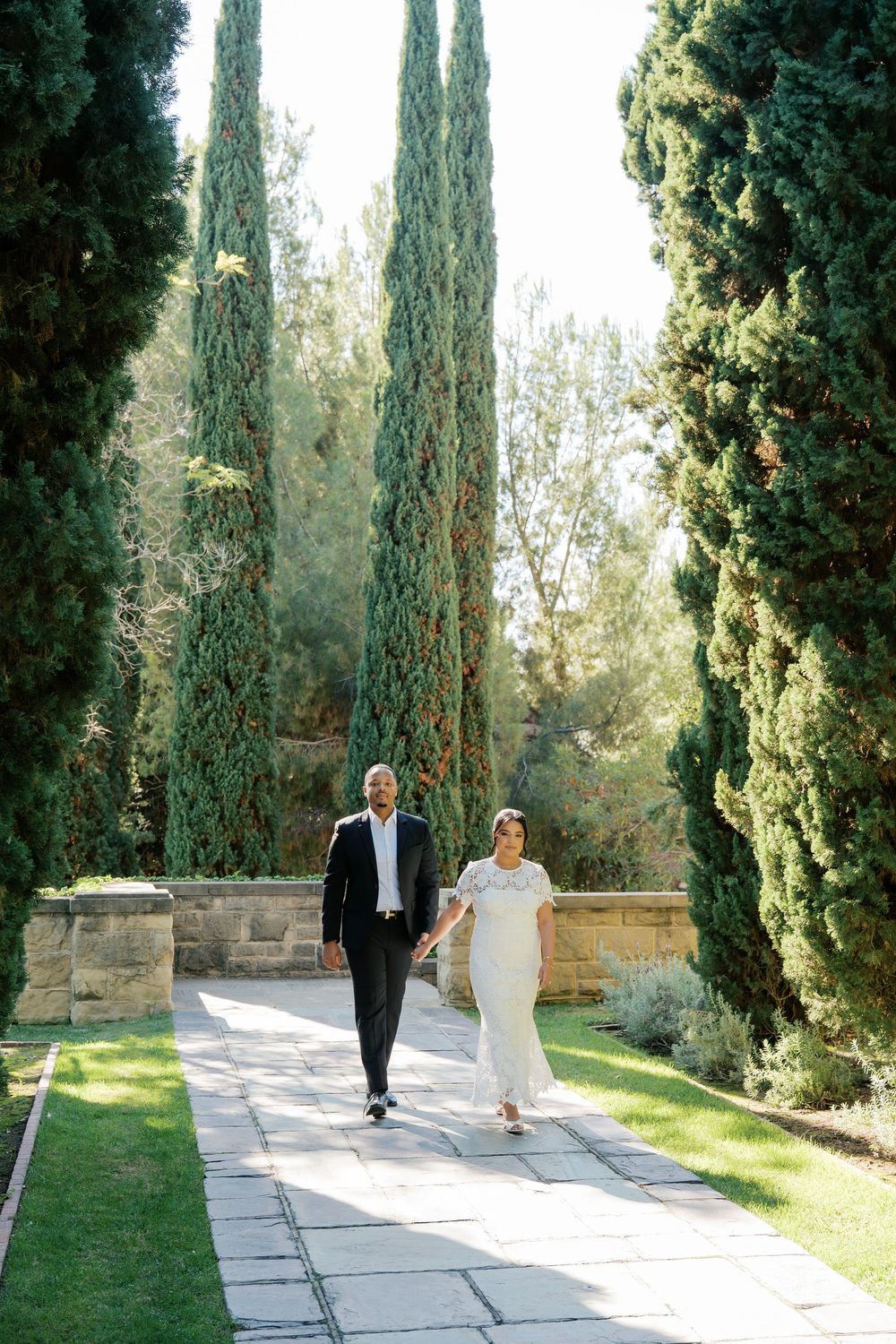 A couple walks along a stone pathway lined with tall cypress trees in an elegant garden setting.