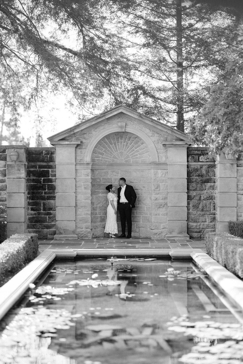Black and white panoramic photograph shows historic stone wall with decorative arched niches and pillars in a garden setting.