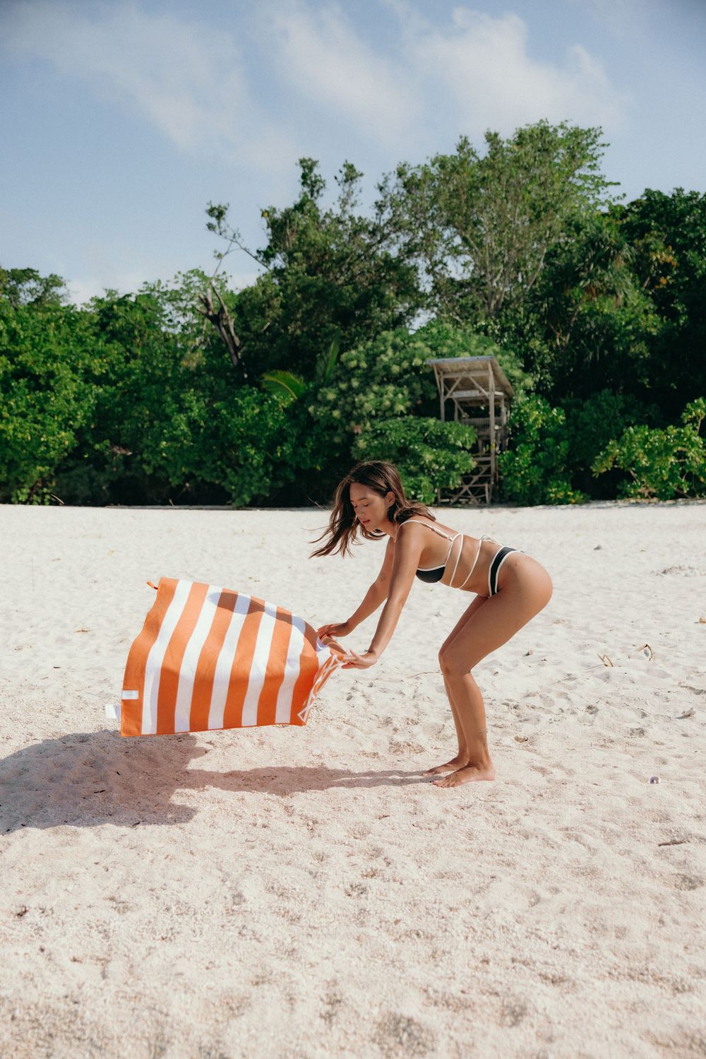 Beach scene with person adjusting striped orange and white beach bag on sandy shore.
