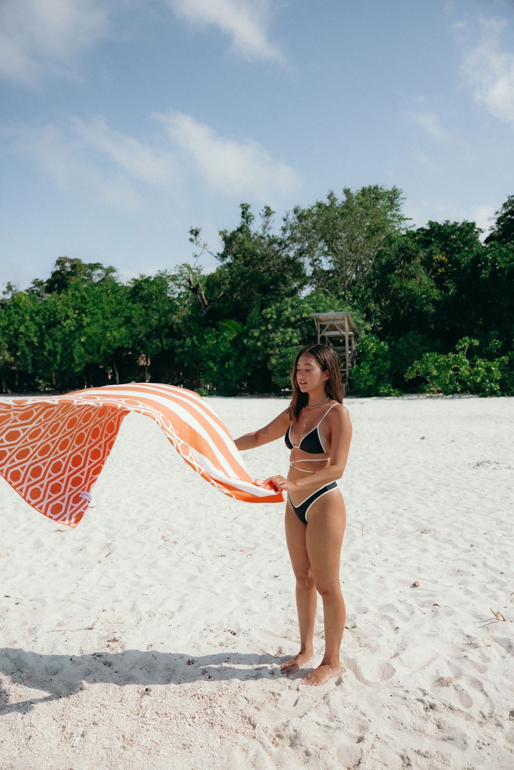 A person waves a bright orange patterned towel on a pristine white sand beach with lush vegetation.