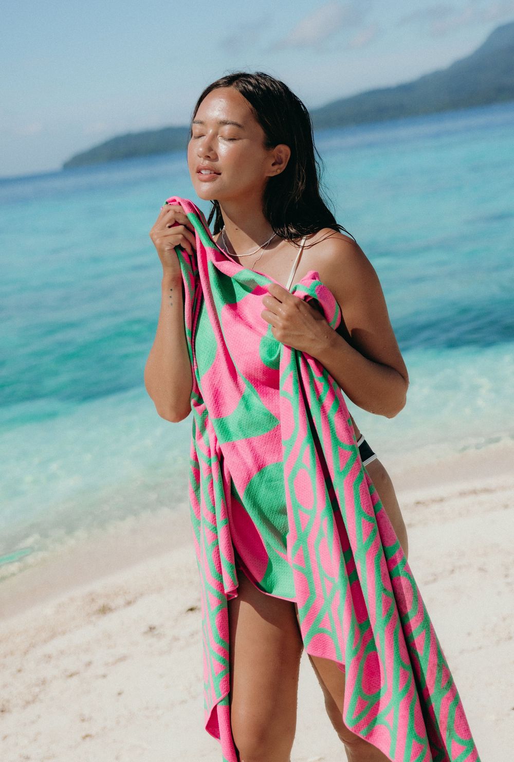 A person in swimwear arranges a vibrant pink and green patterned beach towel by the turquoise ocean.