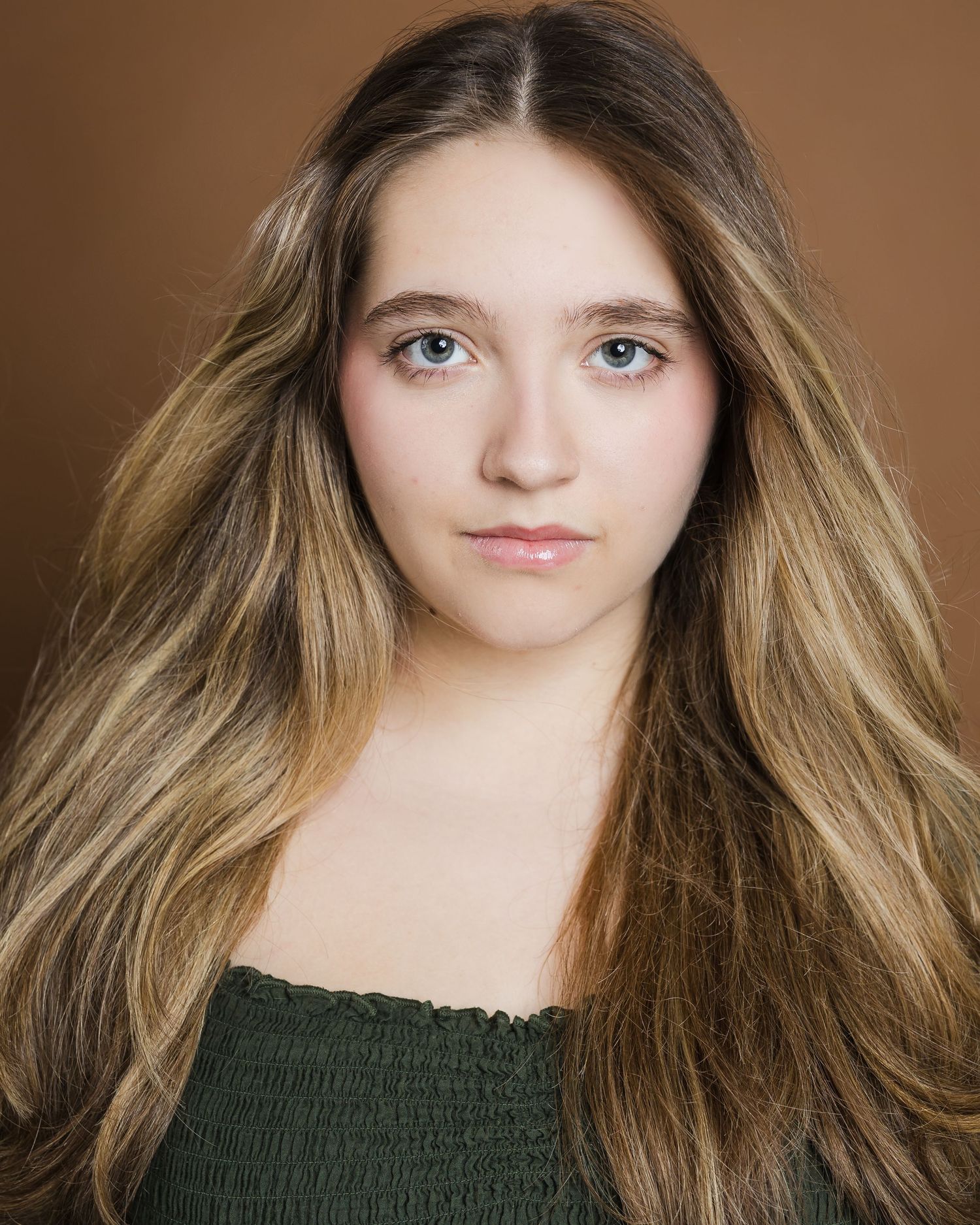 Portrait of person with long wavy brown and blonde hair wearing a dark green top against brown background.