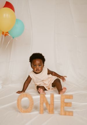 Baby in light colored dress lies on white backdrop with colorful balloons and 'ONE' text display for first birthday shoot.