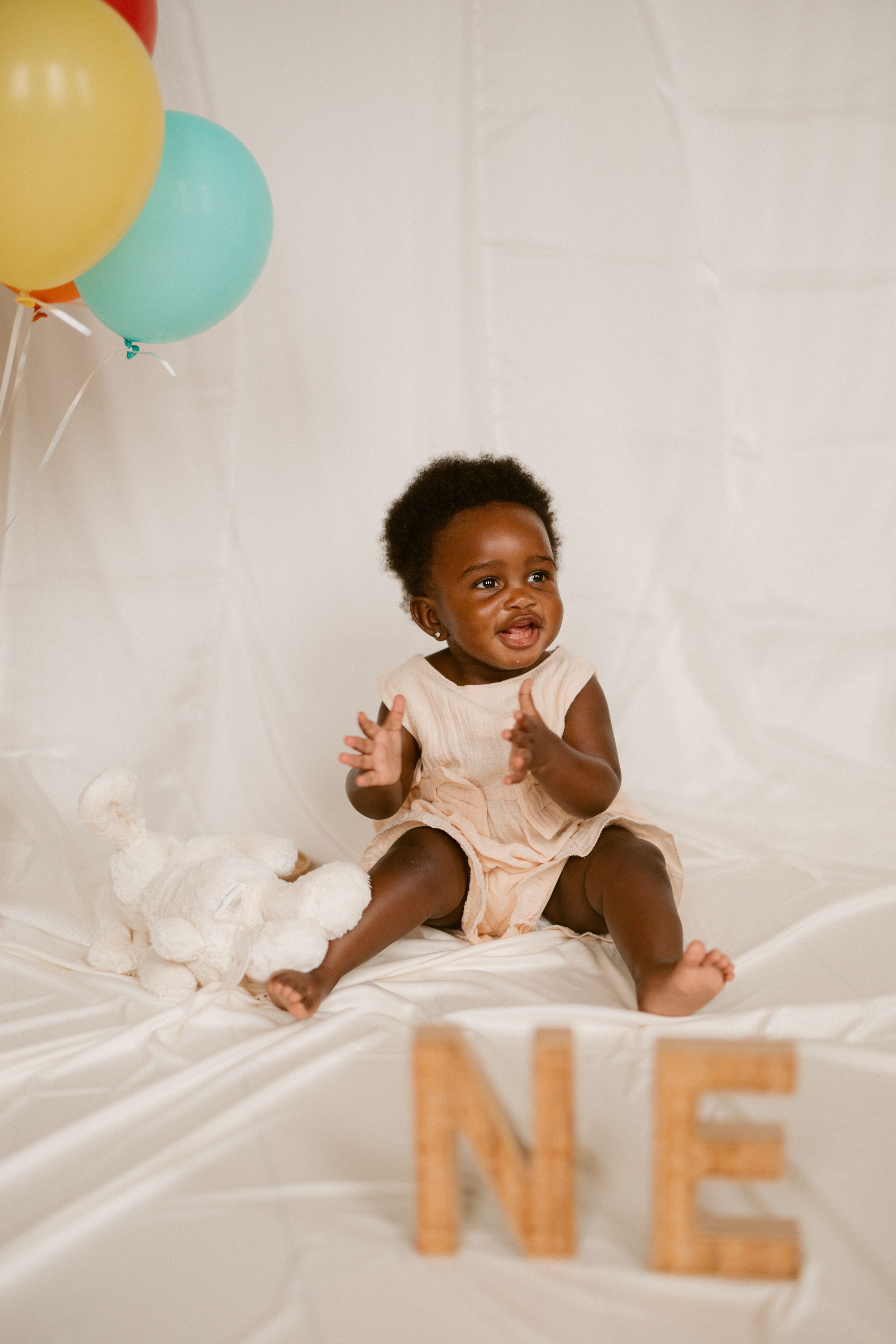 A baby wearing a pink outfit lies on white bedding while colorful balloons float overhead during a photoshoot.