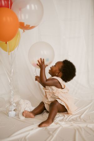 Joyful child plays with colorful balloons while lying on white bedding in a bright studio setting.