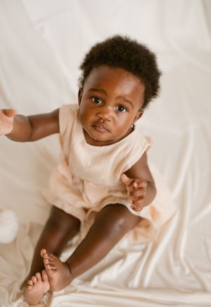 A baby wearing a light colored onesie lies on white bedding while making playful expressions.