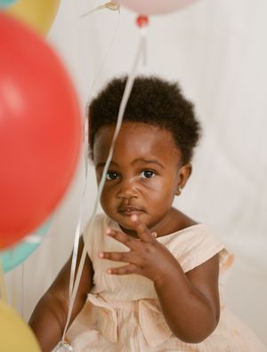 Baby wearing pink dress sits among colorful balloons during birthday celebration photoshoot.