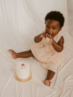 Small child in peach dress plays with cake while sitting on white fabric during photo session.