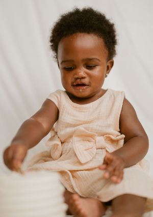 Baby wearing a peach-colored dress with bow detail sits on white bedding while playing and smiling.