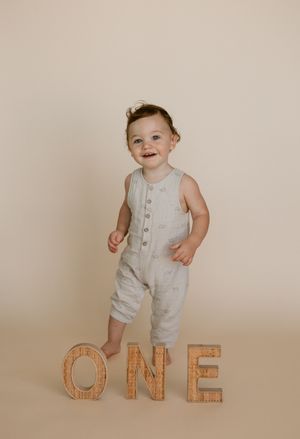 A baby in a light colored romper stands next to wooden block letters spelling 'ONE' against a neutral background.