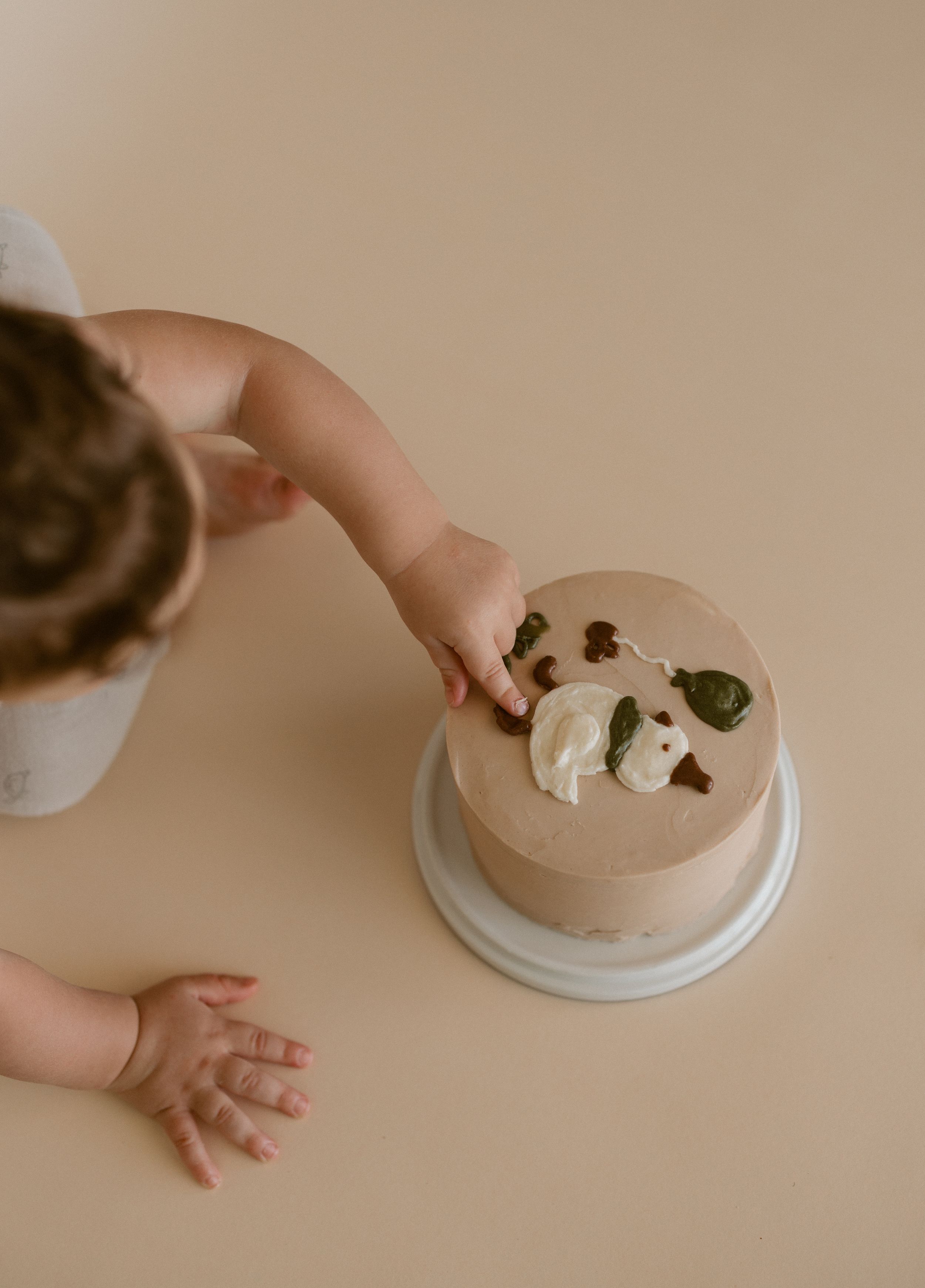 A sequence of hands decorating a light brown mousse or dessert with toppings on a white plate against a neutral background.