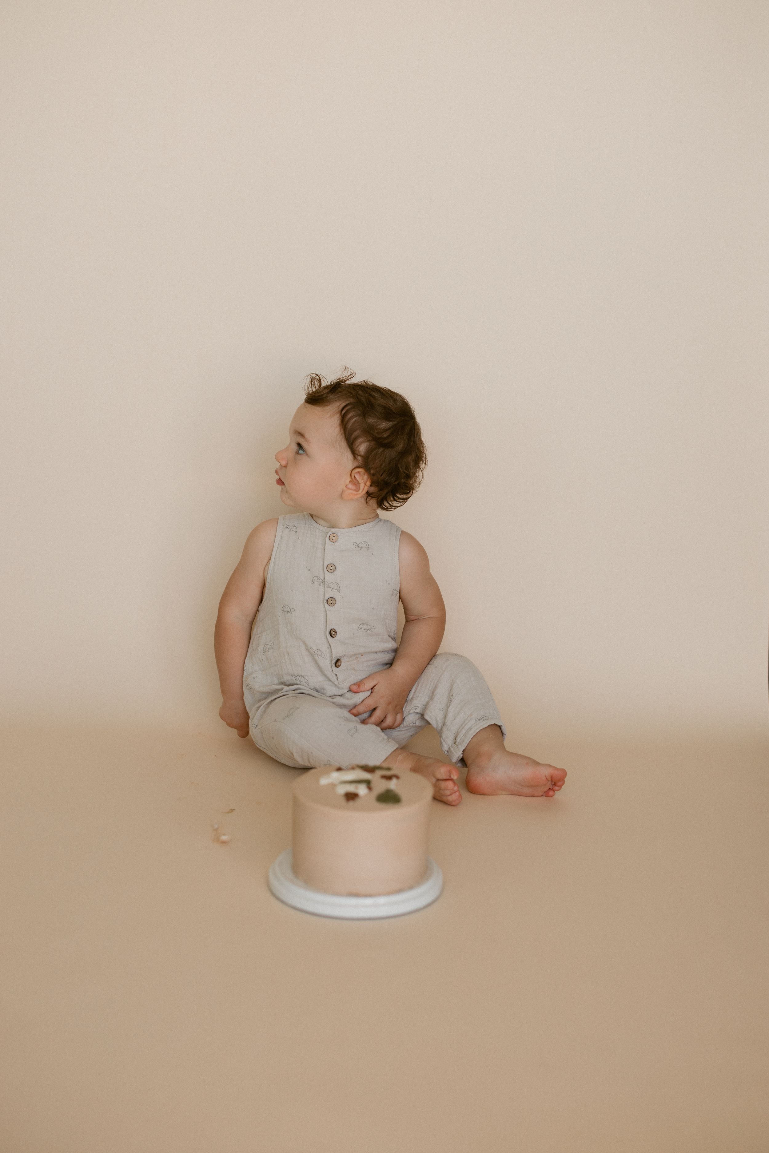 A baby in a gray romper sits and looks around while positioned against a neutral beige background in a series of photos.
