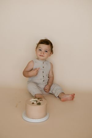 Baby in sleeveless gray outfit sits beside white cake plate on beige background.