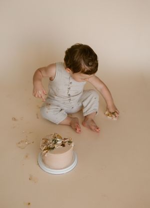 Baby in gray outfit explores and plays with cake crumbs scattered on beige surface.