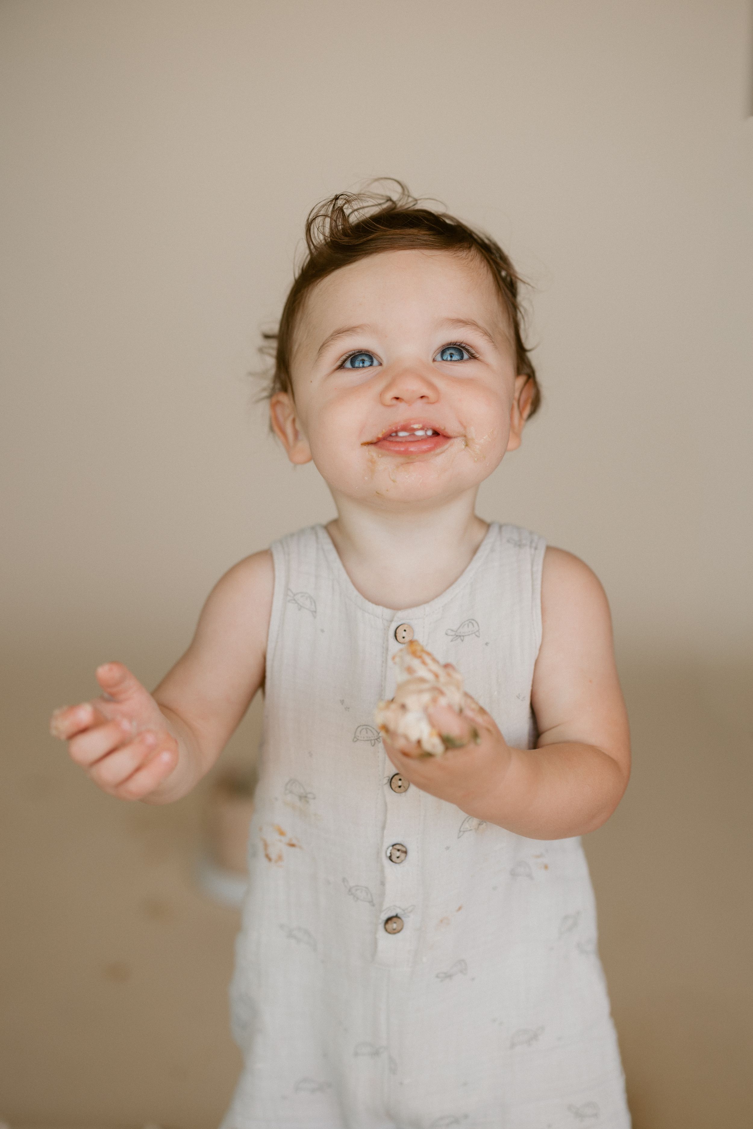 A baby wearing a white sleeveless romper smiles and gestures while sitting against a beige backdrop.