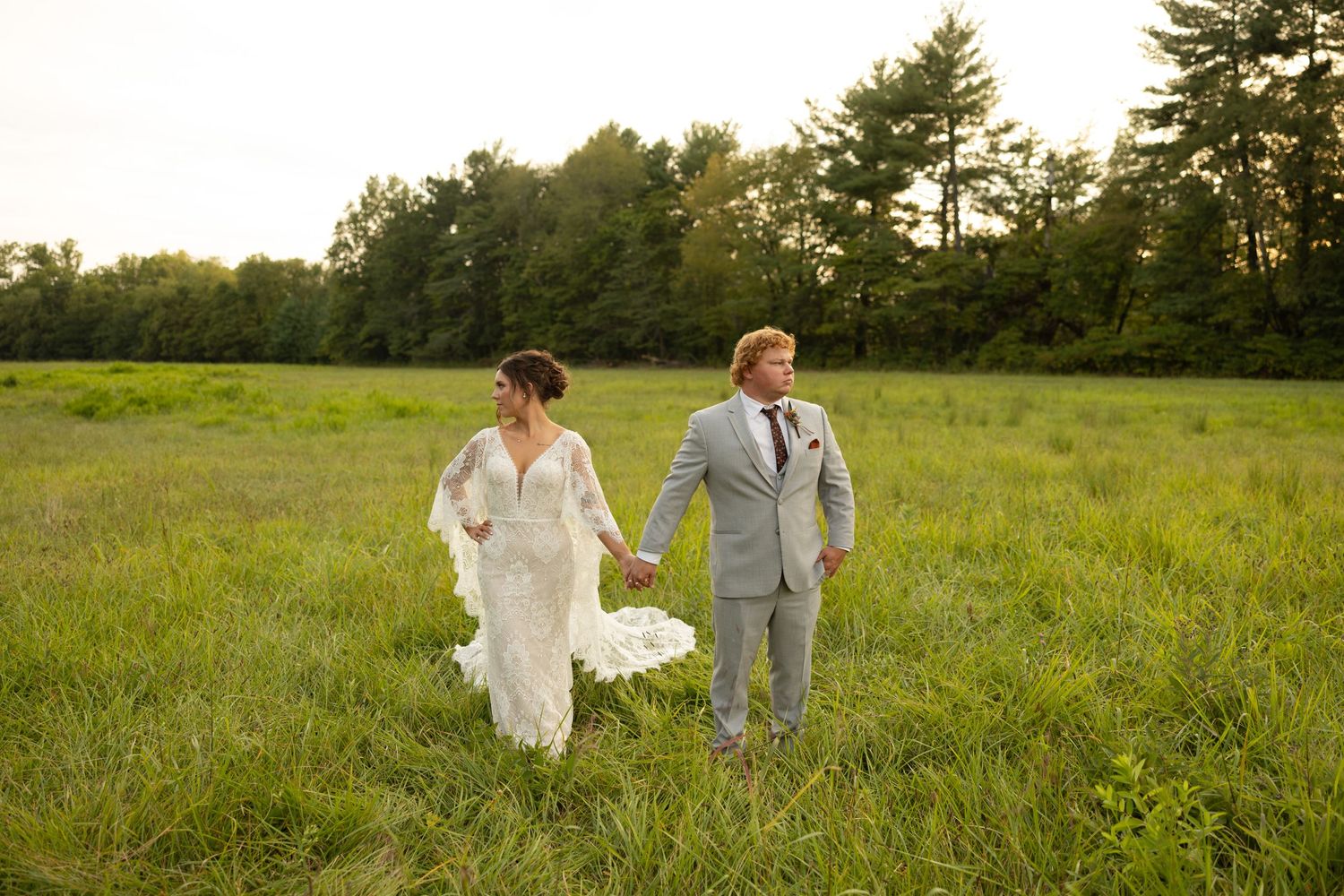 A couple in wedding attire walking hand in hand through a sunlit meadow surrounded by pine trees.