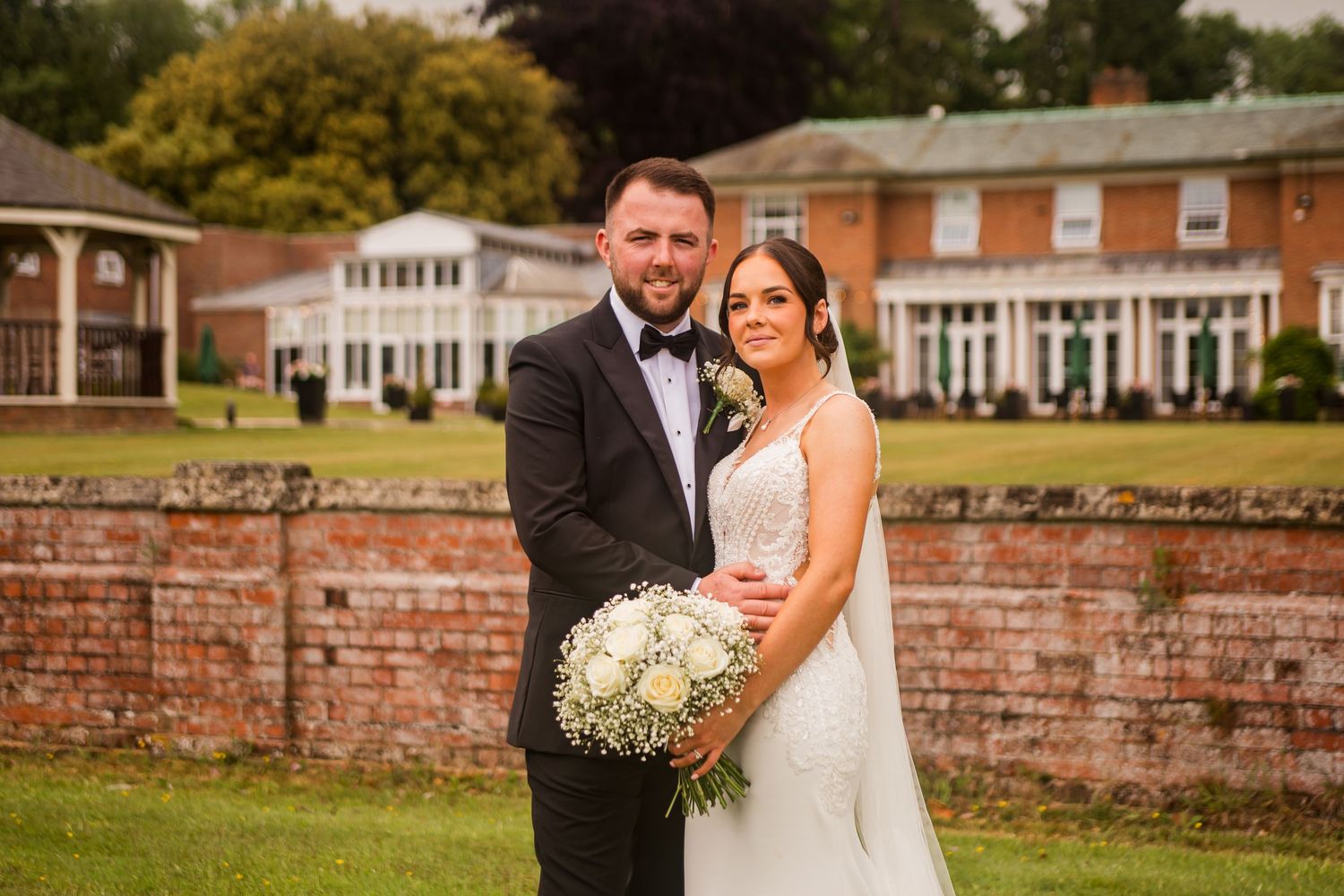 Couple poses for wedding portraits in front of a stately brick mansion with manicured grounds.