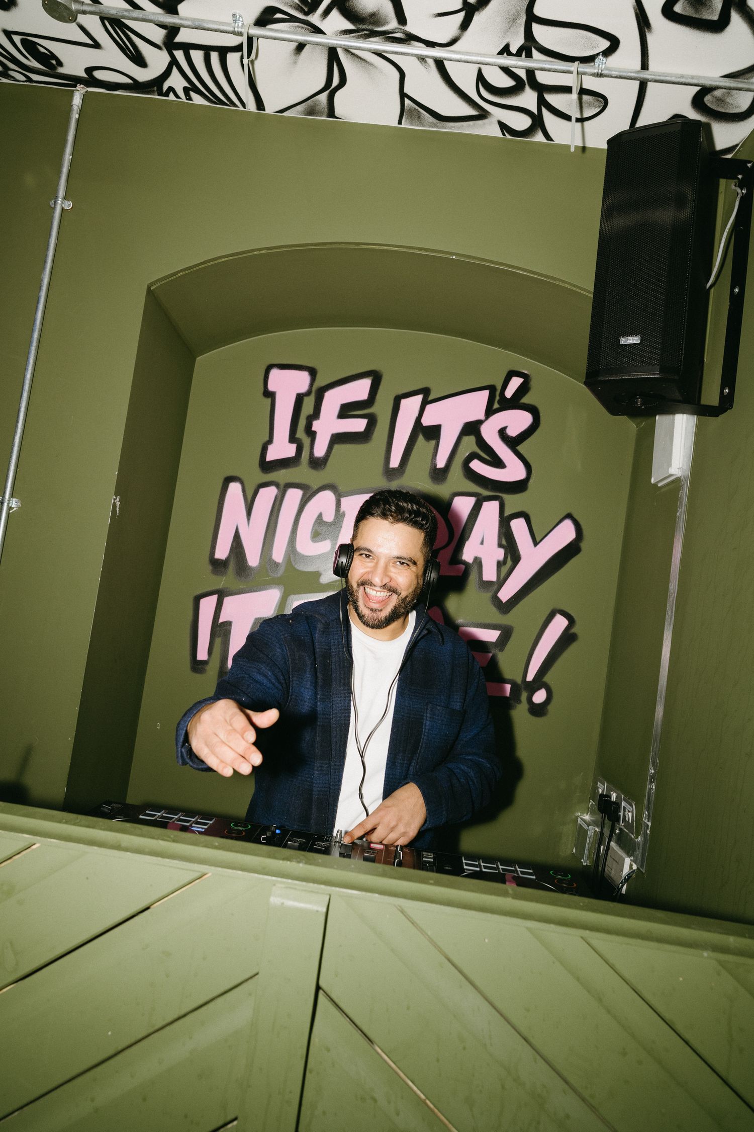 A DJ performs behind decks at a nightclub with pink neon text on the wall behind them.