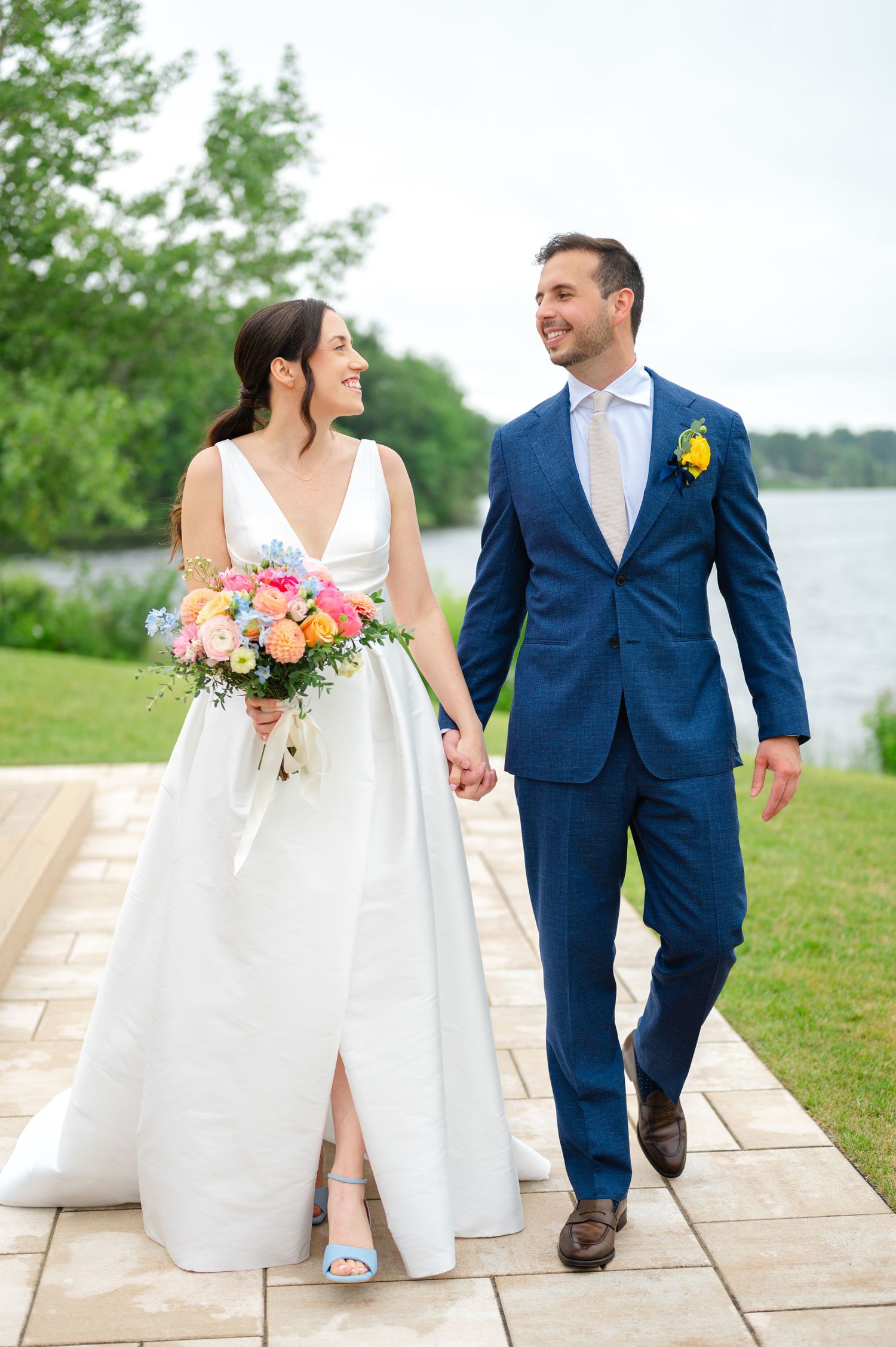 A couple in wedding attire walks hand in hand along a waterfront stone pathway with white chairs and colorful bouquet details.