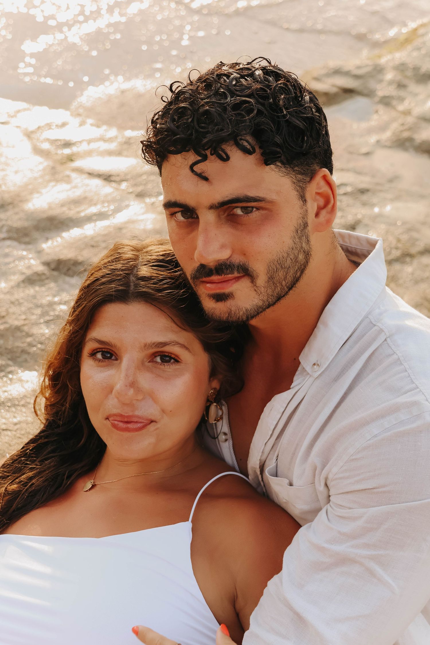 Close portrait of a couple leaning together near sparkling water, man with curly hair in white shirt.