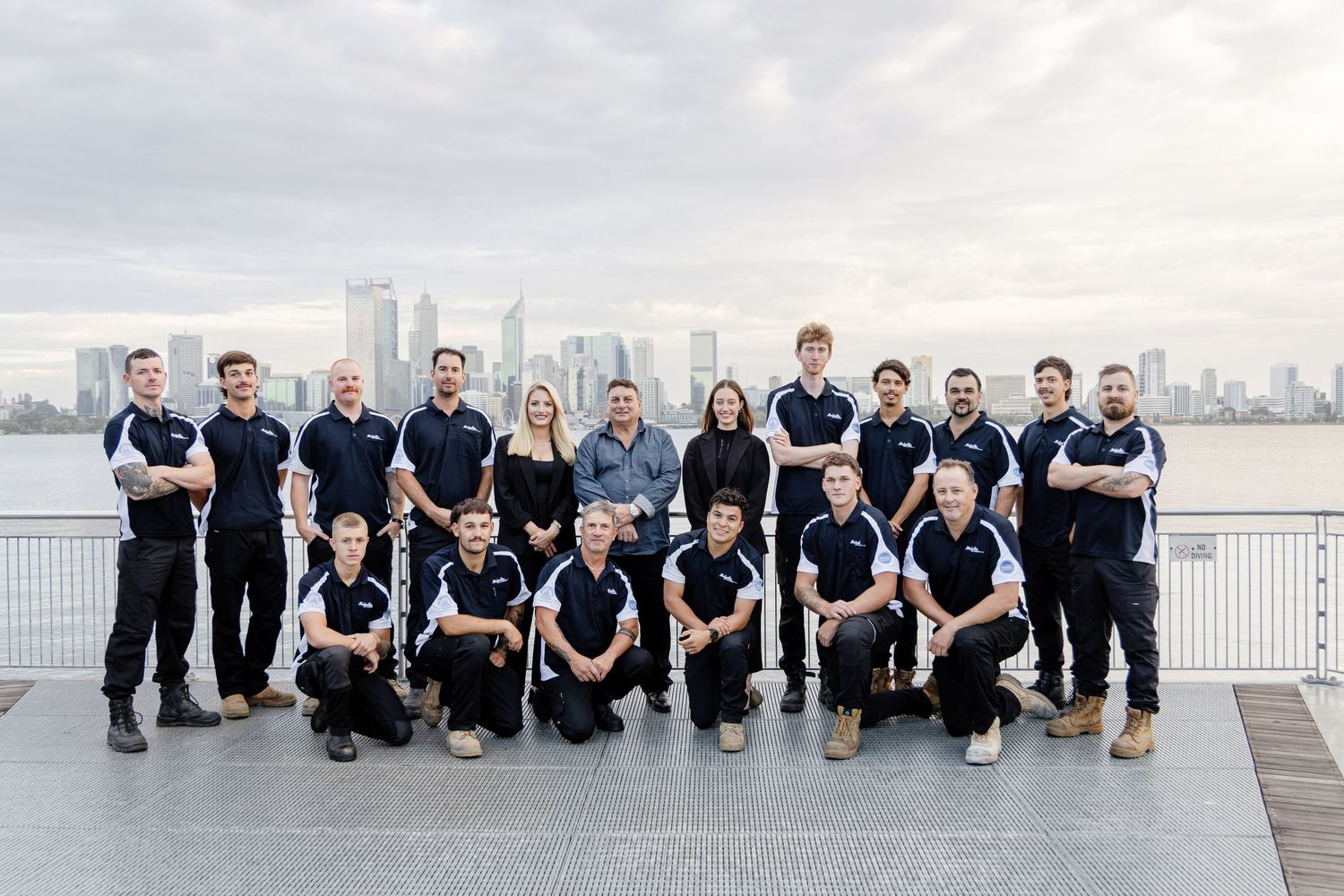 A group in matching navy blue shirts sits together on a white railing overlooking a waterfront cityscape.