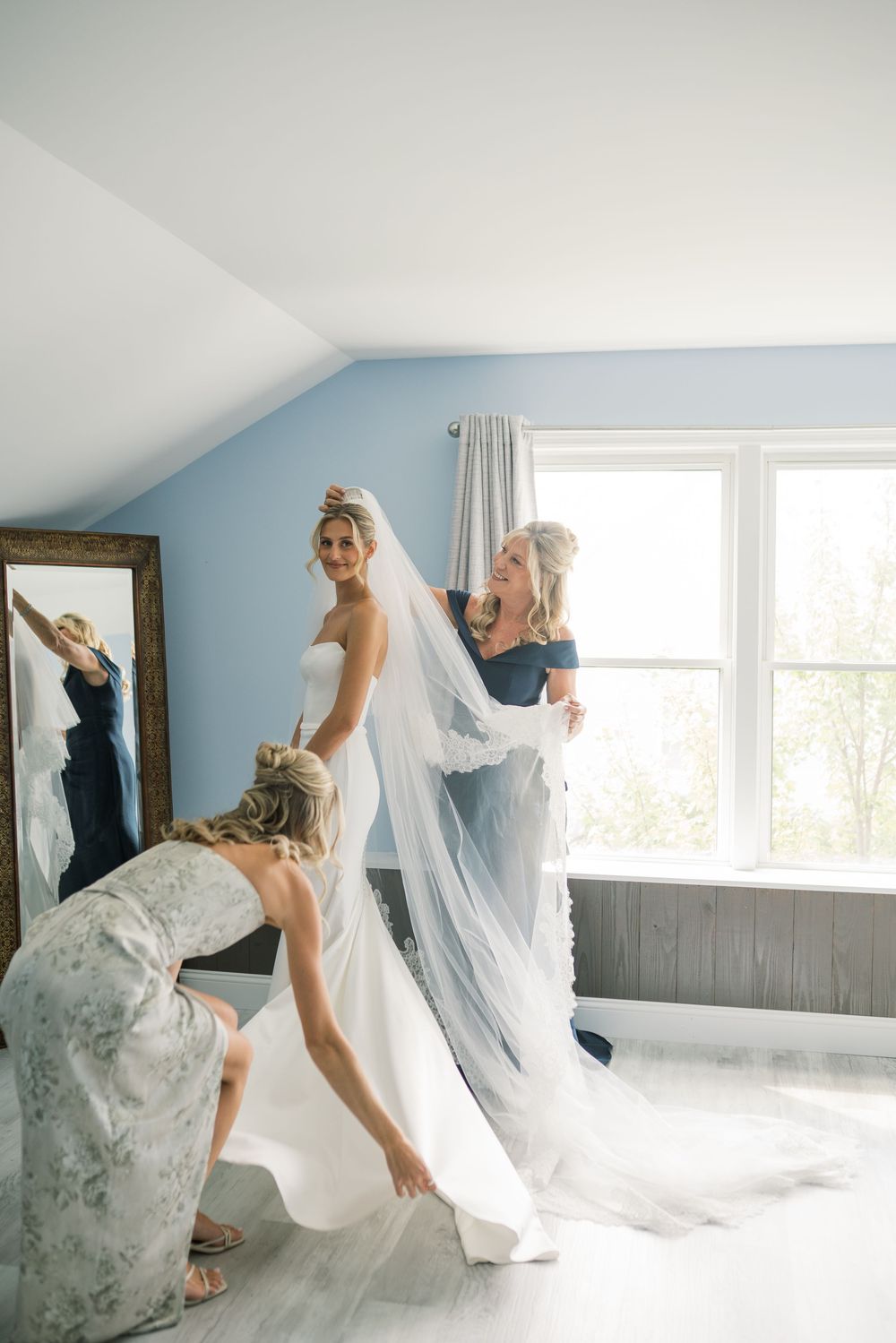 Wedding dress fitting moment in a sunlit room with flowing veil and blue walls.