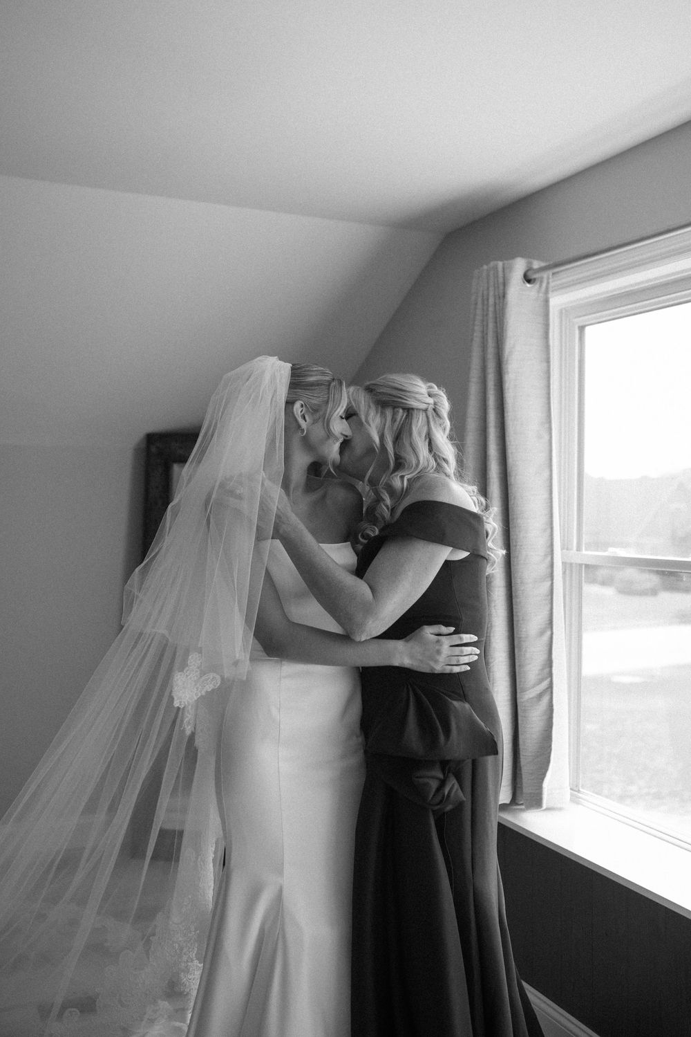 Black and white wedding photograph showing bride in flowing veil embracing groom by a window in an emotional moment.
