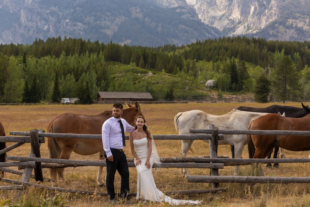 Rustic wooden fences line a path with majestic mountain peaks and evergreen forests in the background.
