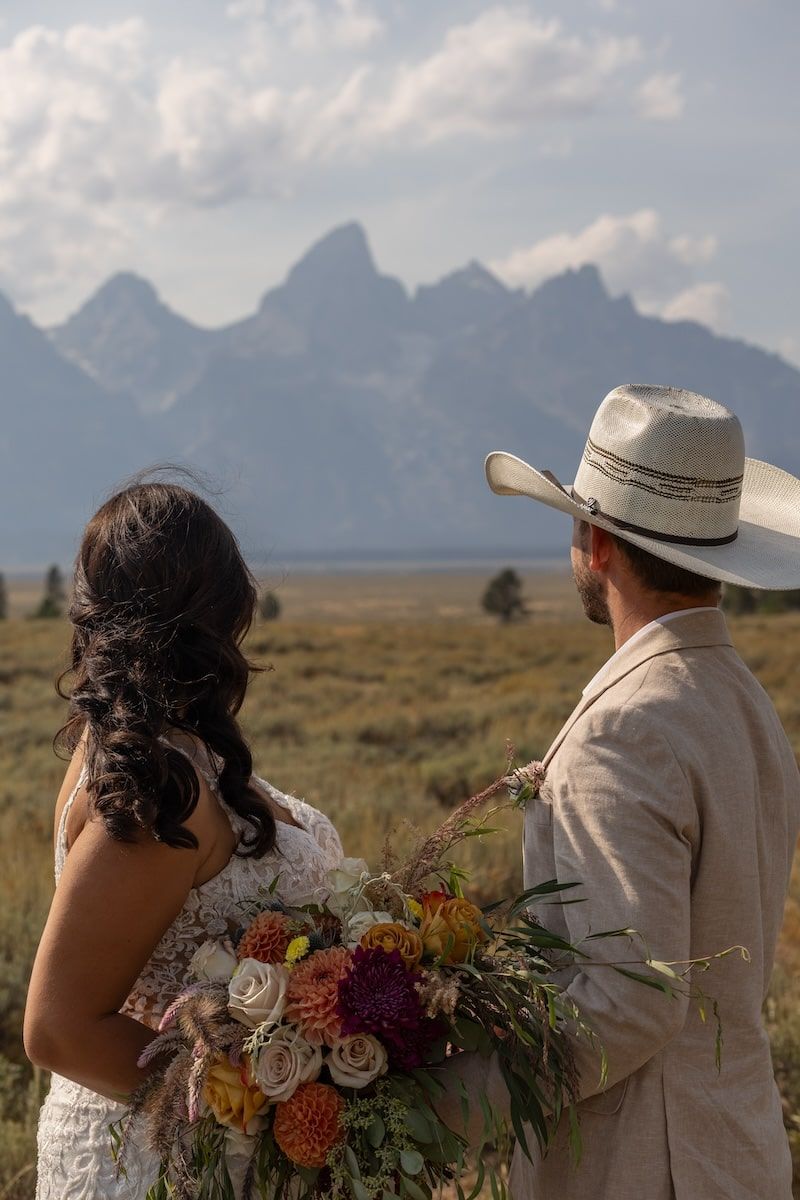 A couple in wedding attire stands together against a backdrop of majestic mountain peaks at sunset.
