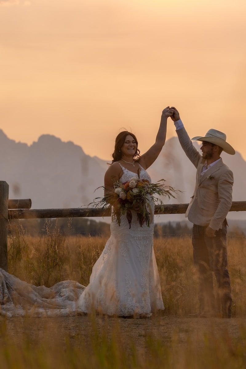 A romantic sunset wedding photo of a couple celebrating with raised hands in a field with mountains in the background.