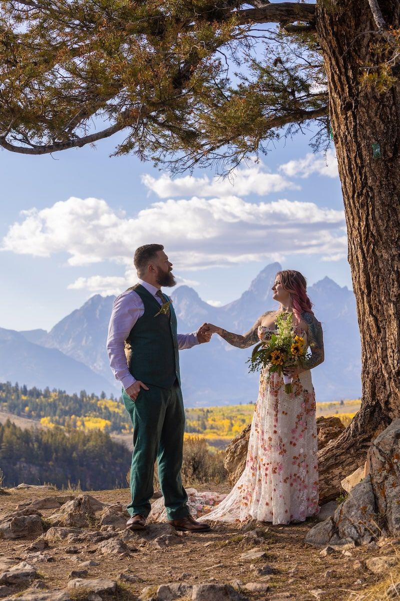 A couple shares a romantic moment under trees with the majestic Grand Teton mountains in the background.