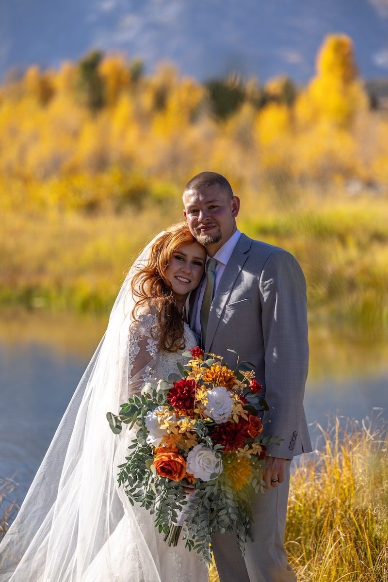 A couple in wedding attire embraces outdoors with autumn foliage and a lake in the background.