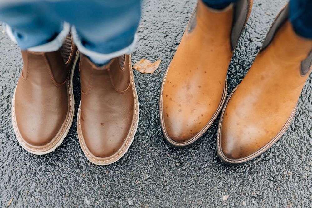 Two pairs of tan leather boots worn by a mother and child standing on asphalt in autumn.