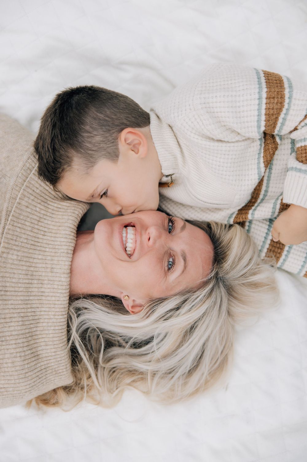 Young boy kissing his laughing blonde mother on the cheek while lying on white bedding.