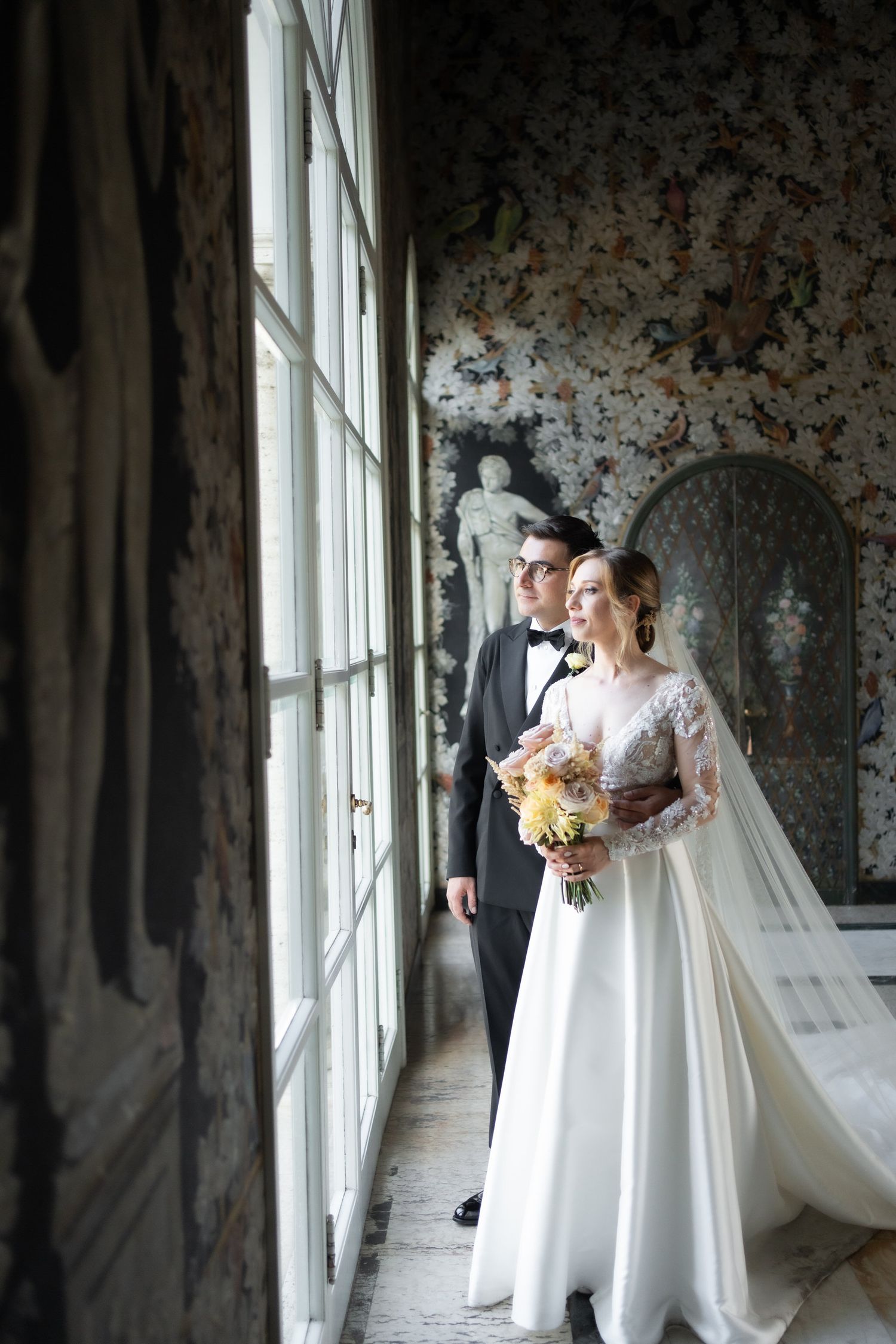 Wedding couple shares romantic moments in a bright hallway with floral wallpaper and large windows.