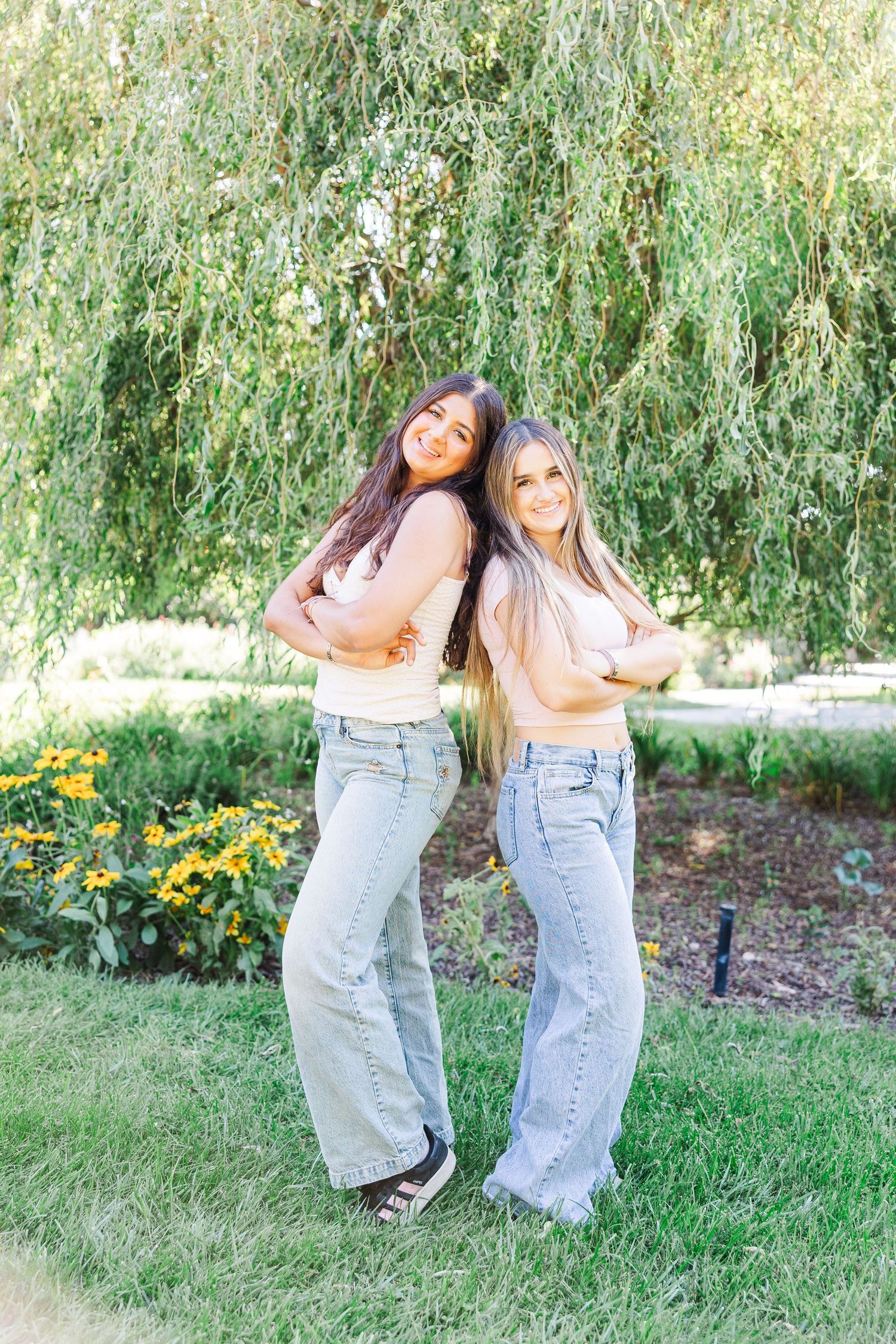 Two friends in matching denim overalls dance and laugh together under a weeping willow tree in a garden setting.