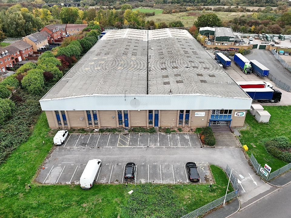 Aerial view of a commercial building with a large empty parking lot surrounded by green landscaping and trees.