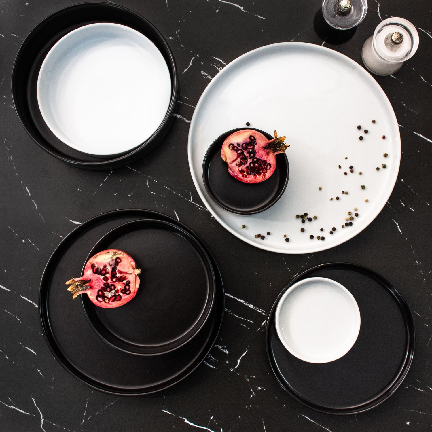 Black and white ceramic plates with pomegranate garnish on dark marble surface with scattered seeds.