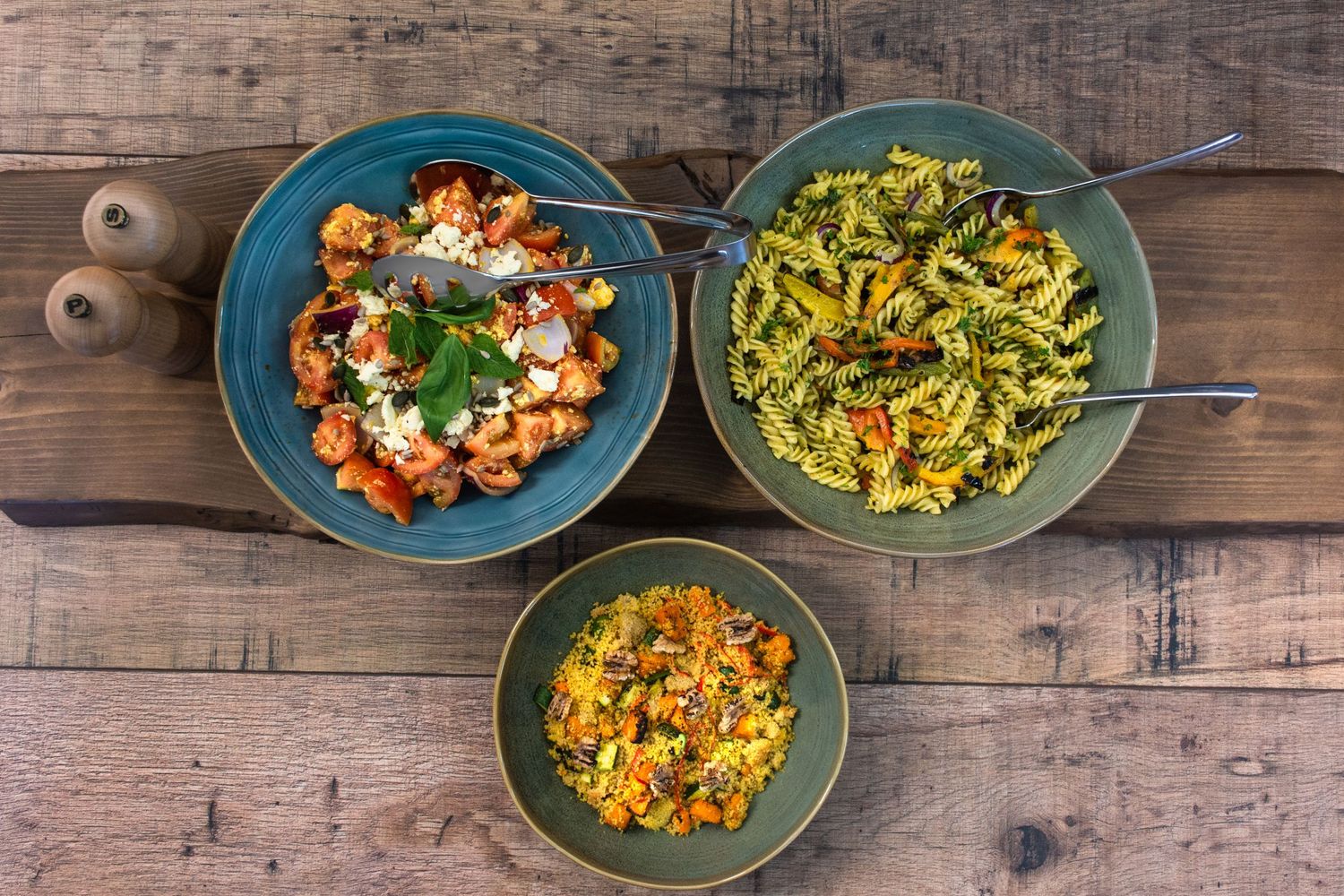 Three bowls of colorful prepared dishes including pasta and salads on rustic wooden table.