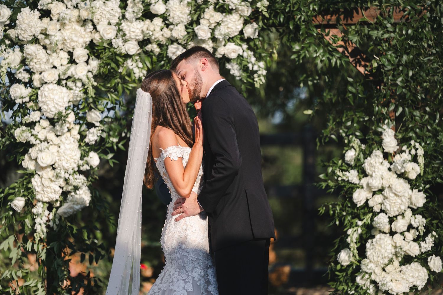 Couple's first kiss with a white roses arch background during their wedding ceremony at The Barn at Villa Venezia NY.
