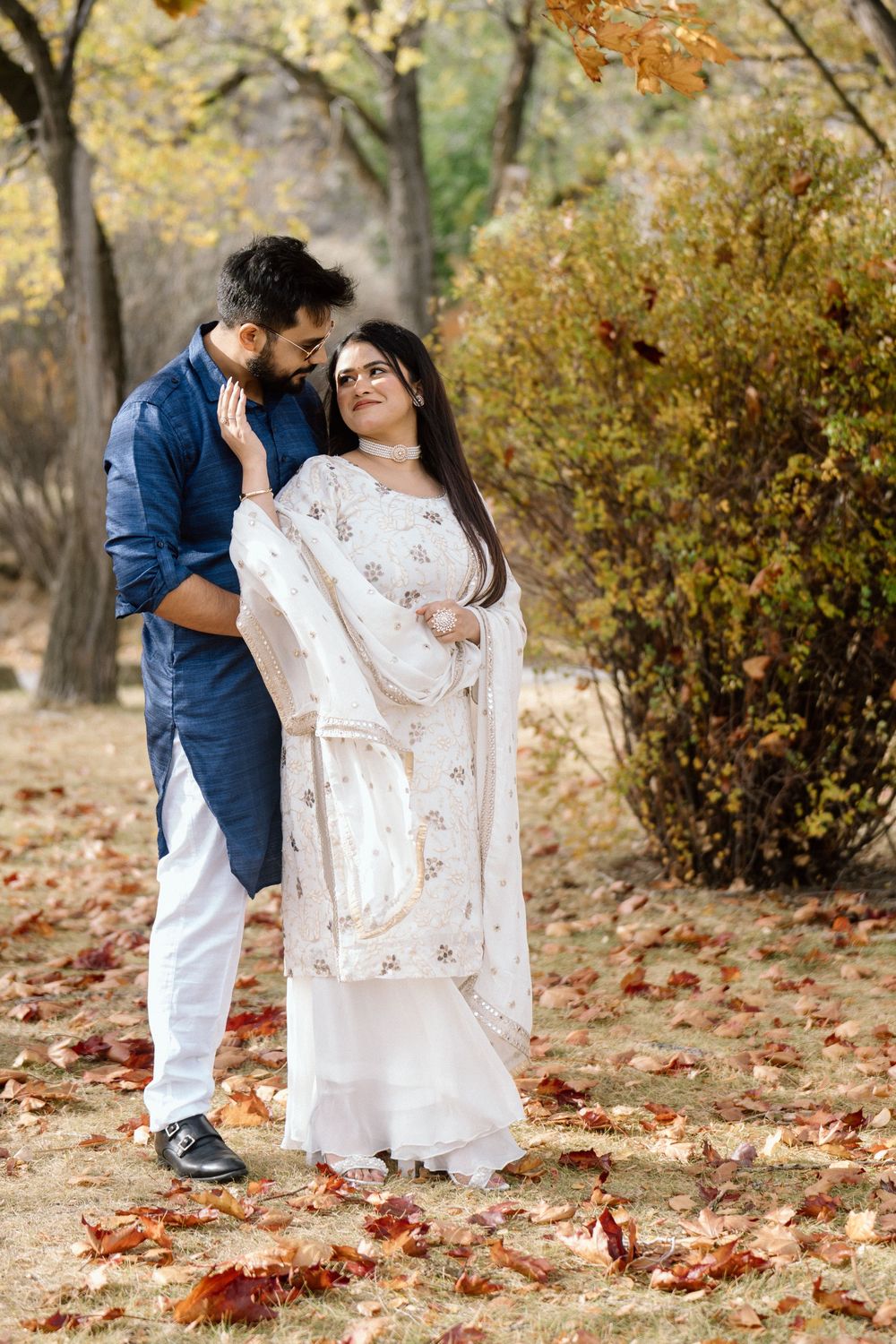 A couple in traditional Indian attire share a romantic moment among autumn leaves in a park setting.