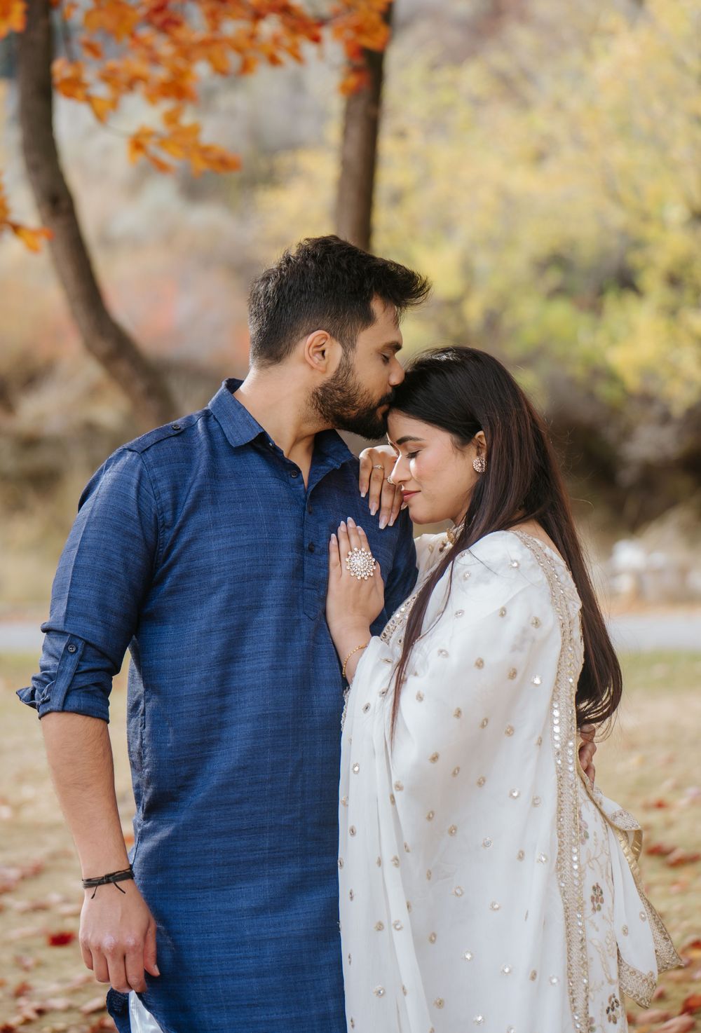 A couple in blue and white outfits share romantic moments amid autumn foliage in a park setting.