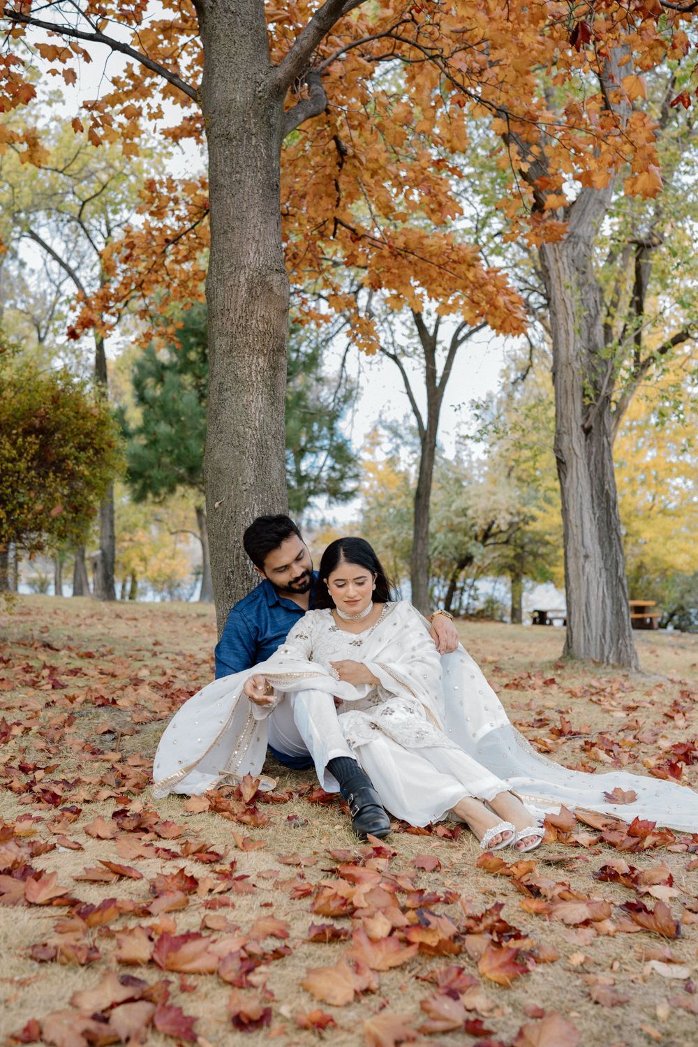 A couple relaxes together on a bed of fallen autumn leaves beneath tall trees in a park setting.