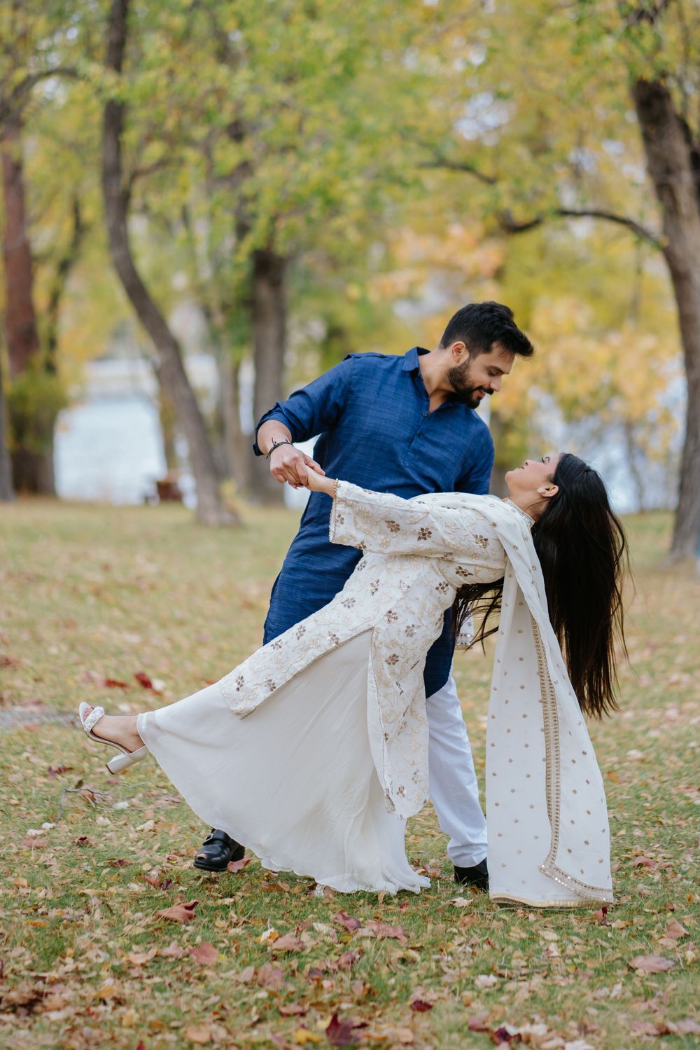 A couple in coordinated blue and white outfits dance romantically among fallen autumn leaves in a park setting.